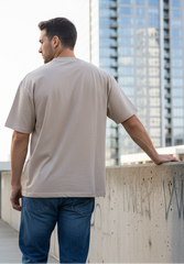 a handsome man wearing plain beige colored oversized t-shirt with blue jeans leaning on wall, showing back side of t-shirt